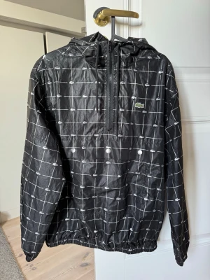 Supreme x Lacoste  - Säljer en svart vindjacka från Supreme x Lacoste med vitt rutmönster och små Lacoste-loggor över hela jackan. Stor Supreme-brodyr på ryggen och klassisk krokodil på bröstet. Jackan har huva, halv dragkedja och elastiska muddar. Perfekt för dig som gillar streetwear.