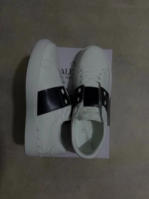 Valentino Garavani vita sneakers - KAN DUSKITERAS OM PRIS! Snygga vita sneakers från Valentino Garavani med bred mörkblå rem över ovansidan och coola nitar längs sulan. Skorna har rund tå, platt sula och klassisk snörning. Märkeslogga i guld på plösen och insidan. Perfekta för dig som gillar exklusiv streetstyle.