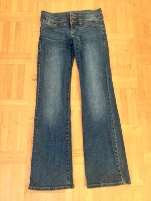 Blå bootcut jeans med låg midja💖 - Säljer ett par blå jeans med bootcut och låg midja. Jeansen har flera knappar i midjan och snygga detaljer baktill med ficklock och knappar. Klassisk tvättad denimlook och raka ben som blir vidare nedtill.