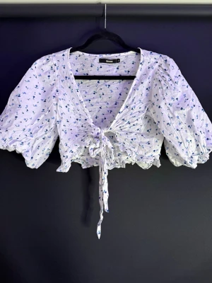 Blommig knytblus från Bik Bok - Supersöt blommig blus från Bik Bok i vitt med små blå blommor. Modellen är croppad med puffiga trekvartsärmar, v-ringning och knyt framtill. Materialet känns lätt och luftigt, perfekt för varma dagar.