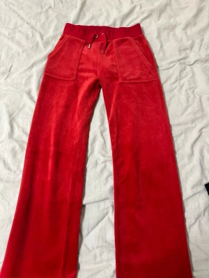 Röda mjukisbyxor Juicy Couture XXS - Säljer ett par klassiska röda mjukisbyxor från Juicy Couture i mjuk velour. Byxorna har bred resårmidja med snörning, raka ben och stora fickor fram med broderad JC-logga. Perfekta för en chill och trendig look. Ena metallbiten på snöret har trillat av se bild 