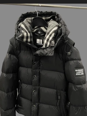 Svart pufferjacka från Burberry - Säljer en svart pufferjacka från Burberry med stor huva och klassiskt rutigt foder. Jackan har dragkedja, knappar och snörning i huvan samt en patch med Burberry London England på ärmen. Perfekt för kalla dagar och riktigt snygg streetstil.