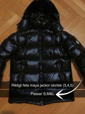 Svart Moncler pufferjacka med huva - Säljer en riktigt fet svart pufferjacka från Moncler med glansig finish och huva. Jackan har dragkedja framtill, två sidofickor och klassisk Moncler-logga på ärmen. Perfekt för kalla dagar när du vill se stilren ut.