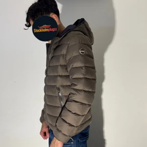 Brun dunjacka från Colmar med huva - Säljer en brun dunjacka från Colmar med huva och två sidofickor med dragkedja. Jackan har ett quiltat mönster och en snygg, normal passform. Perfekt för dig som vill ha en stilren look under höst och vinter.