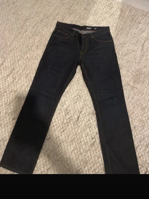 Tommy Hilfiger Mercer Regular Fit Jeans - Säljer ett par mörkblå Tommy Hilfiger Mercer Regular Fit jeans i klassisk rak modell. Jeansen har snygga kontrastsömmar, fem fickor och det ikoniska lilla Hilfiger-flaggan på bakfickan. Tillverkade i slitstarkt jeansmaterial och passar perfekt till en clean och stilren look.