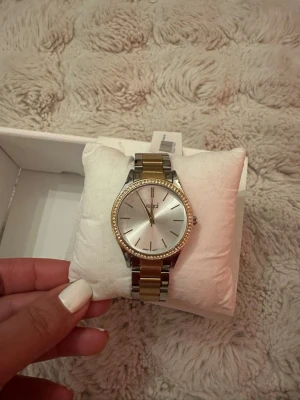 Oanvänd Regal damklocka  - Elegant damklocka från Regal. Klockan har en guldfärgad boett med strassdetaljer runt urtavlan. Armbandet är i tvåfärgat stål, guld och silver. Perfekt accessoar för både vardag och fest. Batteri medföljer inte