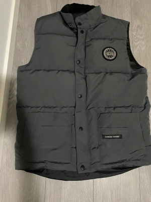 Svart dunväst från Canada Goose M - Säljer en svart dunväst från Canada Goose med hög krage, två stora fickor framtill och klassisk logotyp på bröstet. Västen har tryckknappar och är perfekt för lager-på-lager under kyliga dagar. Enkel och stilren design som funkar till det mesta.