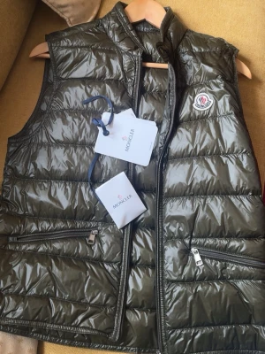 Mörkgrön dunväst från Moncler - Olivgrön gui väst från Moncler,  Storlek 1 som motsvarar xs/s. säljs nu pågrund av att jag behöver kapital till Magaluf 2026🙏, taggen samt extra knapparna till västen finns kvar, mycket bra skick då jag själv knappt använt den😁