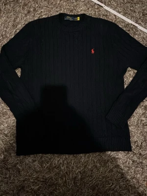 Svart stickad tröja från Polo Ralph Lauren - Säljer en svart stickad tröja från Polo Ralph Lauren med klassisk röd logga broderad på bröstet. Tröjan har rund halsringning, långärmad passform och ribbade muddar. Perfekt för lager-på-lager under kyliga dagar.