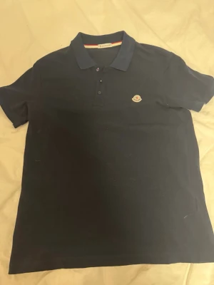 Mörkblå pikétröja från Moncler - Snygg mörkblå pikétröja från Moncler med klassisk krage och tre knappar framtill. Tröjan har korta ärmar och en liten broderad Moncler-logga på bröstet. Perfekt till jeans eller shorts för en clean look.