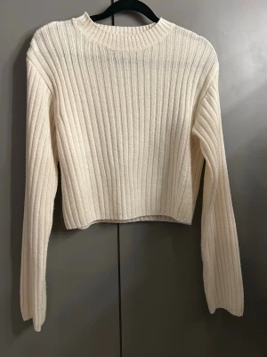 Ribbad vit stickad tröja - Säljer en ribbad vit stickad tröja med lång ärm och rund hals. Tröjan har en croppad passform och passar perfekt om man har ett längre linne under.