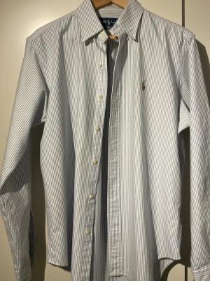 Randig skjorta från Ralph Lauren M - Klassisk skjorta från Ralph Lauren i blåvitrandigt med broderad logga på bröstet. Skjortan har lång ärm, knappar framtill och en classic fit. Skjortan är i mycket fint skick.