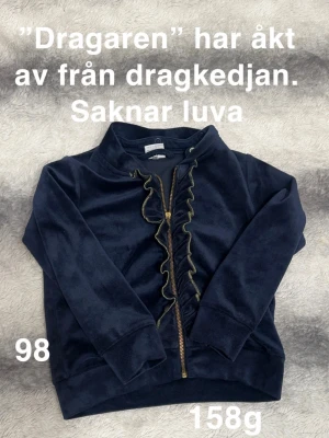 Mörkblå velourkofta med volang - Mysig mörkblå kofta i mjuk velour från Polarn o pyret. Dragkedja framtill med dekorativ volang längs öppningen och guldfärgade detaljer. Långärmad och utan huva, perfekt för lager på lager under kyliga dagar.🌸 Dragaren” har åkt av från dragkedjan. Saknar luva. Storlek 98