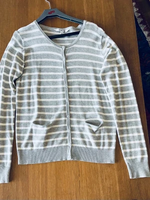 Gråvit randig kofta från Basic U - Säljer en grå och vit randig kofta från Basic U i storlek M. Koftan har långa ärmar, rund halsringning och knäppning framtill med matchande knappar. Tillverkad i mjuk viskosmix och har två små fickor framtill. Perfekmt för lager på lager under kyliga dagar.