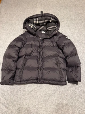 Svart dunjacka från Burberry - Säljer en svart pufferjacka från Burberry med avtagbara ärmar så den kan användas som väst. Jackan har huva med klassiskt rutigt foder, dragkedja och knappar framtill samt två fickor med dragkedja. Märke på ärmen och riktigt schysst passform.