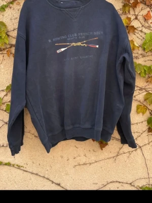  Yves Saint Laurent crewneck - Säljer en mörkblå crewneck från Yves Saint Laurent med tryck av roddklubbs-tema och texten 'R. Rowing Club French Sect.' på bröstet. Tröjan har rund halsringning, långa ärmar och är gjord i mjuk bomull. Perfekt för dig som gillar  klassisk stil.