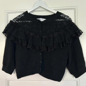 Svart kofta med volang från Zara - Säljer en svart kofta från Zara med snygga volanger och spetsdetaljer över axlarna. Koftan har knappar framtill och är stickad med trekvartsärm. Perfekt för dig som gillar unika detaljer och vill ha en cool look.🖤🖤