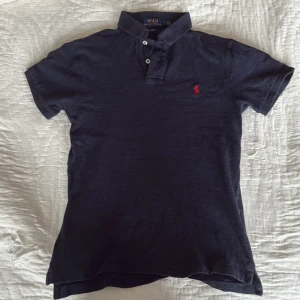 Mörkblå Polo Ralph Lauren Pike - Snygg mörkblå Piké från Polo Ralph Lauren i slim fit med klassisk krage och tre knappar framtill. Ikonisk röd logga broderad på bröstet. Tillverkad i mjuk bomull, perfekt för en clean och stilren look. Sitter mer som en storlek S