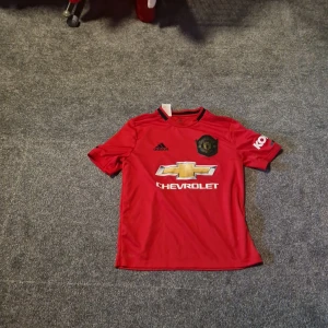 Manchester United röd Adidas matchtröja - Säljer en officiell Manchester United matchtröja från Adidas i rött med svarta detaljer. Tröjan har korta ärmar, klubbmärke på bröstet och Chevrolet-logga i guld och vitt framtill. Materialet är lätt och andas, perfekt för fotbollsträning eller match.