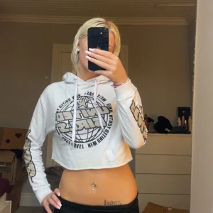 Vit croppad hoodie från Divided XS - Säljer en vit croppad hoodie från Divided med trycket 'United Girls' i glitter på bröstet och ärmarna. Tröjan har huva med snörning och långa ärmar. Perfekt för dig som gillar streetstyle och vill sticka ut lite extra.