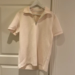 Beige mönstrad t-shirt med krage - Snygg beige t-shirt med korta ärmar och coolt geometriskt mönster i strukturen. Har en krage och halv dragkedja framtill som ger en sportig vibe. Perfekt för dig som gillar stilrena men unika plagg.