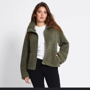 Olivgrön teddyjacka med dragkedja - Mysig olivgrön teddyjacka med hög krage och hel dragkedja framtill. Jackan har två stora fickor och en avslappnad passform som ger en chill vibe. Perfekt för lager-på-lager och att hålla sig varm under kyliga dagar. Använd fåtal ggr och inga skador. Som ny! Storlek S men passar mig som har xs 