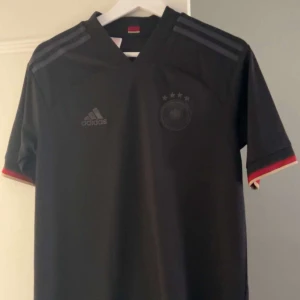 Svart Tysklands fotbollströja Adidas - Snygg svart Tysklands fotbollströja från Adidas med diskret logga och emblem på bröstet. Tröjan har korta ärmar med röd och gul detalj vid mudden samt klassiska Adidas-ränder på axlarna. Tillverkad i lätt och ventilerande polyester, perfekt för match eller träning.