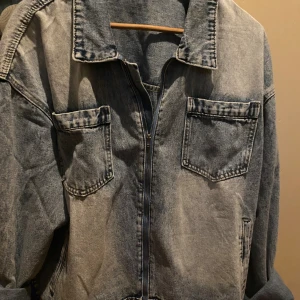 Grå jeansjacka med dragkedja - Snygg grå jeansjacka med dragkedja framtill och två stora bröstfickor. Jackan har klassisk krage och raka ärmar, perfekt för en avslappnad streetstil. Materialet är denim och färgen är tvättad grå med en cool vintagekänsla.