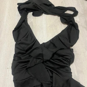 Svart omlottklänning med volang - Snygg svart omlottklänning med volangdetaljer och djup v-ringning. Klänningen har breda axelband och är tillverkad i ett mjukt, följsamt tyg som ger en elegant look.