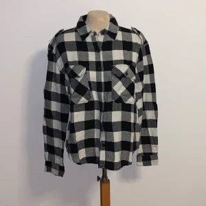 Rutig overshirt från Forever 21 - Snygg svartvit rutig overshirt från Forever 21 med klassisk krage, två stora bröstfickor med lock och knappar. Skjortan är långärmad och har axelklaffar för en cool touch. Perfekt att bära öppen eller stängd över en t-shirt.