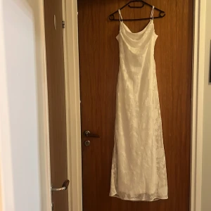 Vit långklänning från Gina - Supersnygg vit långklänning från Gina Tricot med tunna axelband och draperad halsringning. Klänningen har ett subtilt blommönster i tyget som ger en romantisk vibe. Materialet känns lätt och har en satinlik finish. Perfekt för dig som vill sticka ut med en elegant look. Hade på mig klänningen en gång.