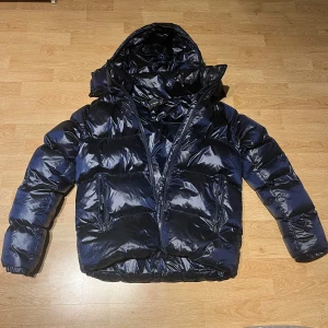 Everest jacka S/M  - Snygg mörkblå pufferjacka med glansig finish och stor huva. Jackan har dragkedja framtill, två fickor med dragkedja och elastiska muddar vid ärmsluten. Perfekt för kalla dagar när du vill hålla värmen och ändå se cool ut.