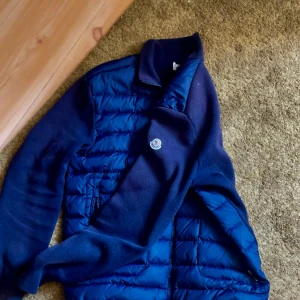 Blå quiltad pufferjacka från Moncler - Snygg marinblå pufferjacka från Moncler med quiltad framsida och ärmar i mjuk ullmix. Jackan har dragkedja framtill, ståkrage och två fickor med dragkedja. Perfekt för dig som vill ha en stilren och sportig look med premiumkänsla.