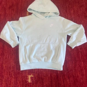 Ljusblå hoodie från Nike - Snygg ljusblå hoodie från Nike med klassisk logga på bröstet. Tröjan har huva, känguruficka och ribbade muddar vid ärmslut och midja. Perfekt för en chill och sportig look.