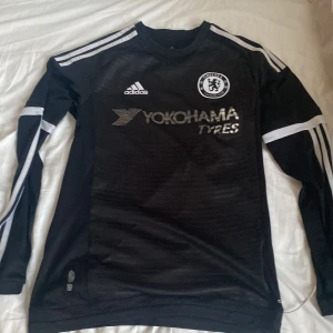 Chelsea långärmad svart Adidas tröja - Chelsea FC långärmad fotbollströja från Adidas i svart med vita detaljer och silverfärgat tryck. Tröjan har rund hals, tre vita ränder på axlar och ärmar, samt Climacool-material som andas. Chelsea-logga och Yokohama Tyres sponsortryck på bröstet.