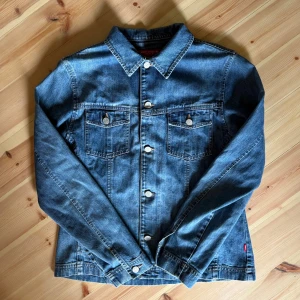 Jeansjacka från Jackpot by Carli Gry - Klassisk blå jeansjacka från Jackpot by Carli Gry. Jackan har raka linjer, två bröstfickor med knappar och silverfärgade knappar framtill. Tillverkad i slitstarkt denimtyg med snygga söm-detaljer och krage. Perfekt för dig som gillar en tidlös och cool stil.