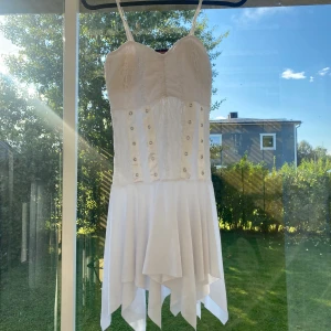 Vit kortklänning med spets och knappar - Säljer en söt vit kortklänning med smala axelband, spetsdetaljer upptill och dekorativa knappar framtill. Kjolen har ett asymmetriskt fall som ger en cool och luftig känsla. Perfekt för varma dagar eller speciella tillfällen!