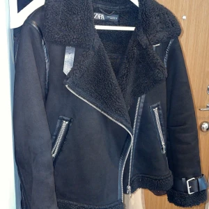 Svart bikerjacka från Zara XS - Säljer en svart biker jacka från Zara i storlek XS. Jackan har stora kragar, dragkedjor på fickor och framtill samt snygga detaljer i skinnimitation. Fodrad med mjuk teddy och har bälte vid nederkanten. 