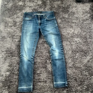 Blå slim jeans från Nudie Jeans - Säljer ett par blå slim jeans från Nudie Jeans med klassisk femficksdesign och snygga slitningar. Jeansen har kontrastsömmar, broderade bakfickor och är tillverkade i stretchig bomullsdenim för en slim passform.