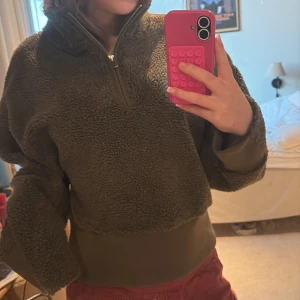 Olivgrön fleece-tröja från MNG - Mysig olivgrön fleece-tröja från MNG med halv dragkedja och hög krage. Tröjan har en loose passform och är perfekt för kyliga dagar. Materialet är fluffigt och mjukt, med breda muddar i midja och ärmslut för extra komfort.