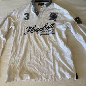 Vit långärmad pikétröja Hackett - Vit långärmad pikétröja från Hackett London med broderad logga, nummer 3 på bröstet och snygga detaljer på kragen. Tailored fit för en schysst passform. Perfekt för dig som gillar sportig och stilren look.