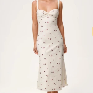 Adoore klänning - Superfin långklänning i ljus beige med små blommiga mönster i rött. Klänningen har omlottbyst, smala axelband och en figurnära passform som ger en snygg siluett. Perfekt för sommarens alla tillfällen och enkel att styla med smycken eller sandaler.