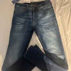 Blå jeans från Jack & Jones - Snygga blå jeans från Jack & Jones med slitningar och lätt tvättad look. Klassisk femficksmodell med gylf och knapp, samt läderpatch bak i midjan. Jeansen har en smal passform och är tillverkade i mjukt bomullsmaterial med lite stretch. W30 L32
