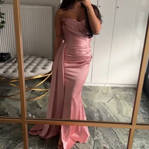 Lång rosa axelbandslös klänning - Säljer en elegant lång klänning i satinliknande material med snygg drapering och omlott-detaljer. Färgen är en mjuk rosa och modellen är axelbandslös med figurnära passform och slits framtill. Perfekt för dig som vill sticka ut med en glamorös look. Fläckar på klänningen se sista bild
