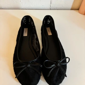 Svarta ballerinaskor med spets från Nelly - Snygga svarta ballerinaskor från Nelly med spetsdetaljer och söt rosett framtill. Skorna har rund tå och är fodrade med ett tunt spetsmönster som ger en extra touch. Perfekta att matcha med både jeans och klänning.