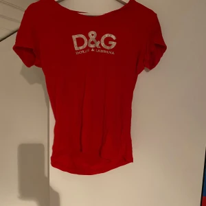 Röd D&G t-shirt med glittertryck - Snygg röd t-shirt från Dolce & Gabbana med korta ärmar och glittrigt D&G-tryck på bröstet. Perfekt för dig som gillar att sticka ut med en ikonisk logga och cool färg. Mjuk och skön bomullskänsla, passar till jeans eller shorts.