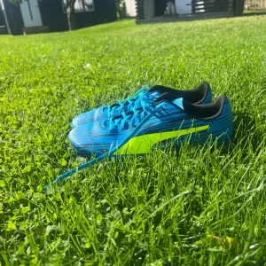 Blåa fotbollsskor från Puma strl 45 - Säljer ett par snygga blåa fotbollsskor från Puma i storlek 45. Skorna har coolt svart mönster, neongul detalj på sidan och svarta innersulor. De har snörning och är tillverkade i syntetmaterial, perfekta för dig som vill sticka ut på planen.