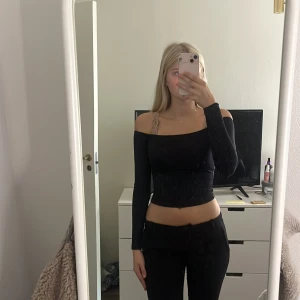 Svart offshoulder topp - Säljer en svart, croppad offshoulder topp med lång ärm. Sitter tight och har en stilren, enkel design utan mönster eller detaljer. Materialet känns mjukt och stretchigt, perfekt för att skapa en clean look. Passar dig som gillar minimalistisk stil.