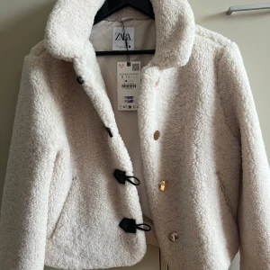 Vit teddyjacka från Zara med detaljer - Supermjuk och fluffig vit teddyjacka från Zara med coola svarta knäppdetaljer och guldiga knappar. Jackan har hög krage, lång ärm och är perfekt för att hålla värmen under kyliga dagar. Snygg och trendig modell som lyfter vilken outfit som helst.