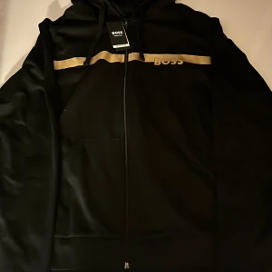 Svart hoodie med guld från BOSS - Svart hoodie från BOSS med dragkedja, huva och snörning. Snygg guldfärgad rand över bröstet och BOSS-logga i guld. Mjuk bomullsblandning och känguruficka framtill. Perfekt för en chill och stilren look.